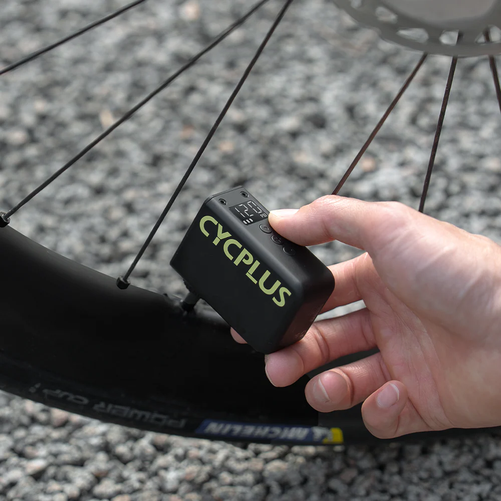 Hand holding a CYCPLUS mini e-pum device on a valve of a bicycle wheel on a road. 