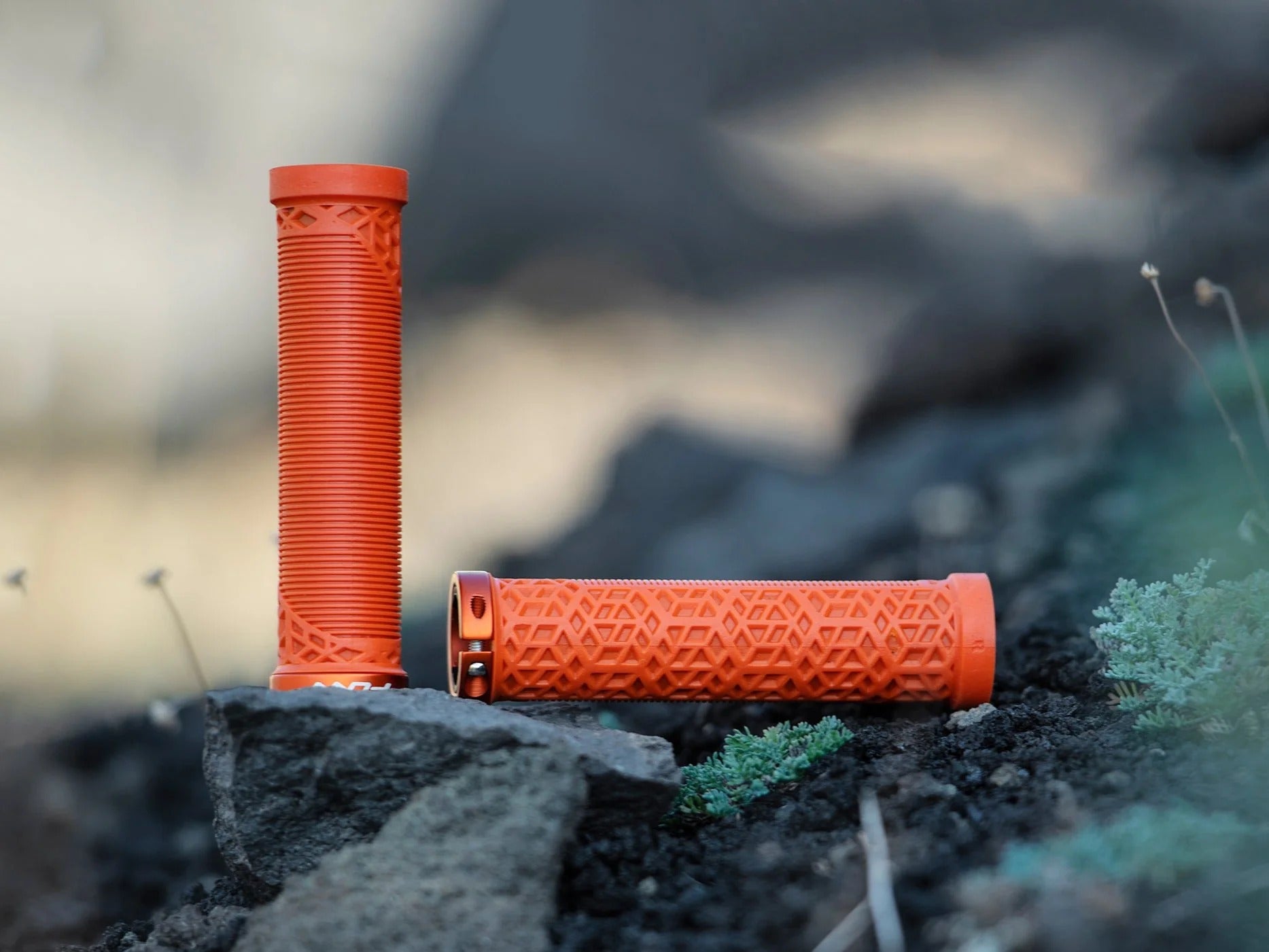 Two bright orange textured bicycle handlebar grips, one standing upright and one lying horizontal, placed on dark rocks with a blurred outdoor background.