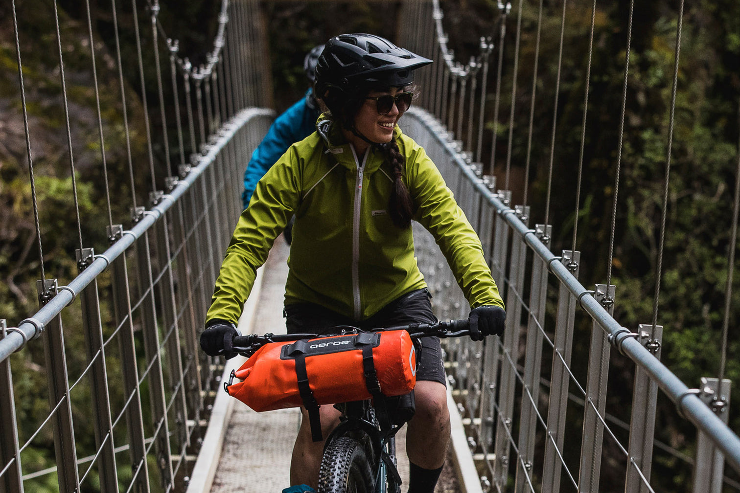 A picture of a girl riding a bike across a bridge with the Aeroe Spider Handlebar Cradle attached to front of bike with a bag