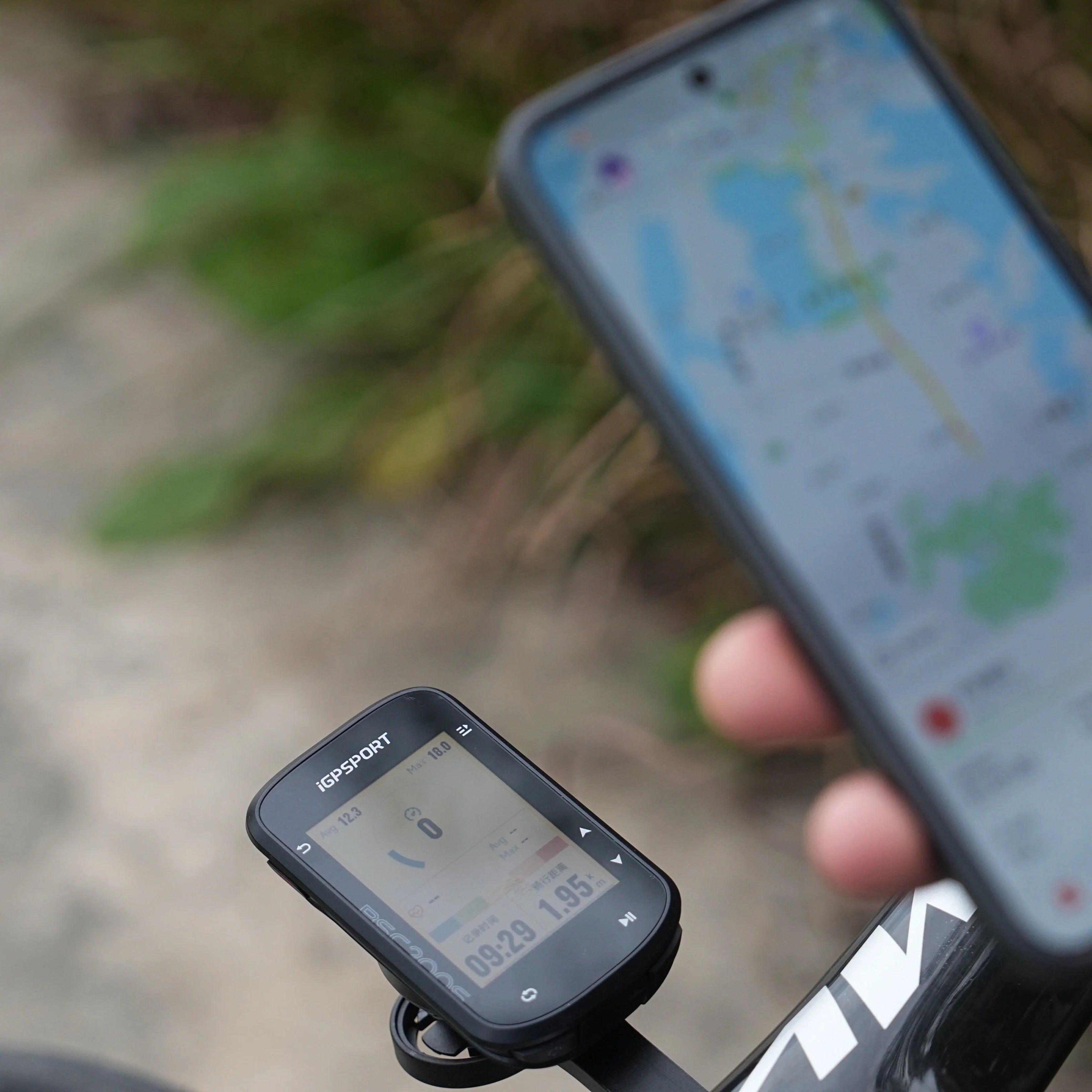 Hand holding a smartphone with a map displayed, next to a cycling computer on a bike handlebar.