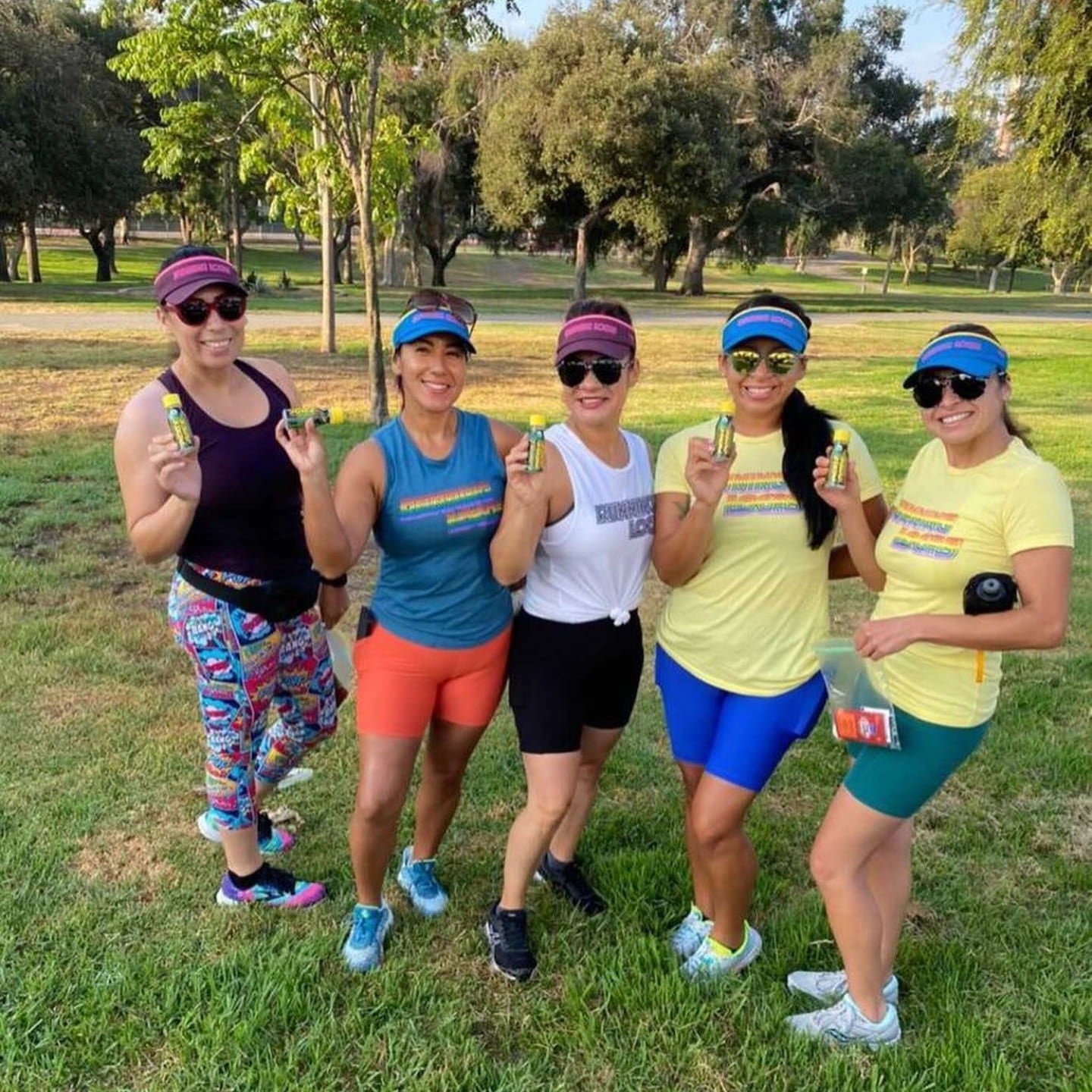 Five women posing outdoors in a park, wearing matching outfits and sunglasses each holding Pickle Juice