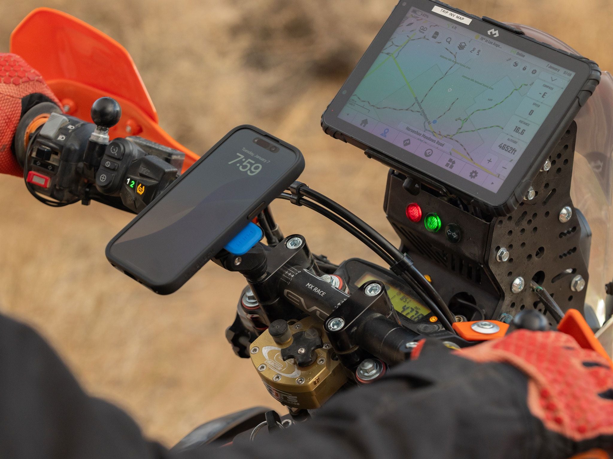 Close-up of a motorcycle handlebar with a GPS device and smartphone mount on a blurred desert background.
