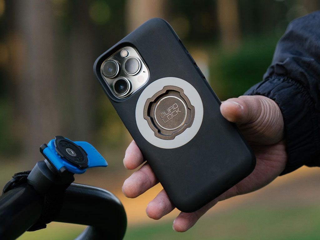 Person holding a black Quad Lock phone case with a ring holder, blurred outdoor background