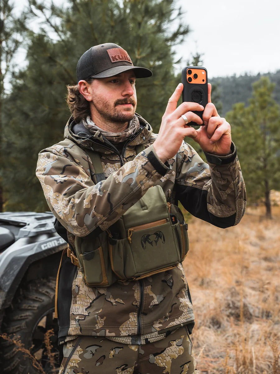 Man in camouflage gear taking a photo with a phone in a forest setting