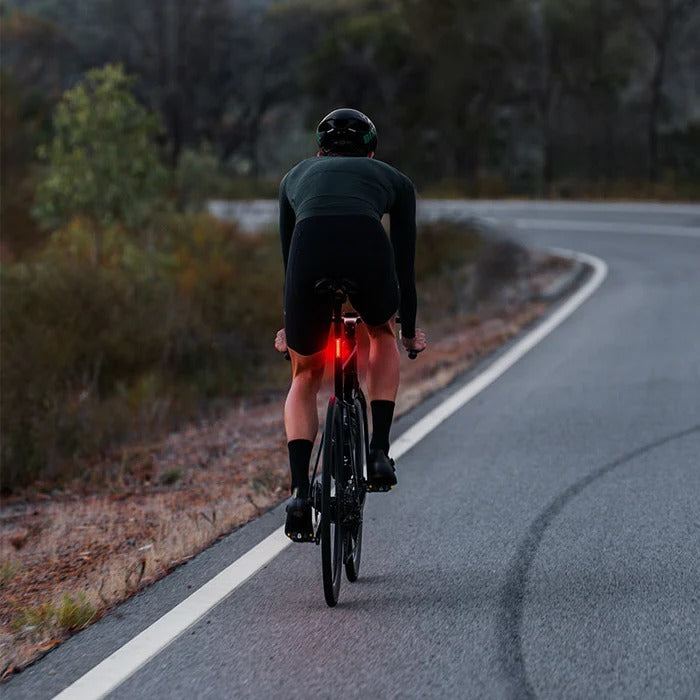 Cyclist riding on a road with a bright red tail light on bike wearing a dark helmet and black outfit, surrounded by greenery.