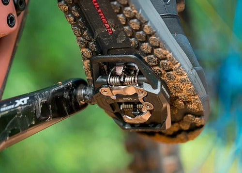 Close up of a shoe clipped into a mountain bike pedal with sand on the bottom of the shoe and a blurry outdoor background