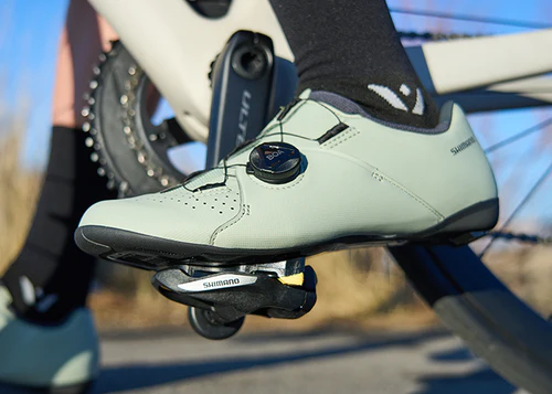 Close-up of a light green cycling shoe clipped into Shimano pedals on a blurred bike background