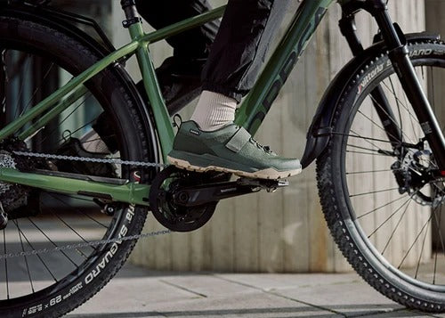 A close-up lifestyle shot of a person riding a green mountain bike, wearing green and white cycling shoes with black pants. The bike frame, tyres, and drivetrain are clearly visible against an outdoor concrete background.
