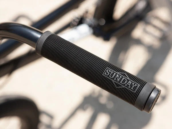 Close-up of a black bicycle handlebar grip with 'Sunday' branding on a blurred background.