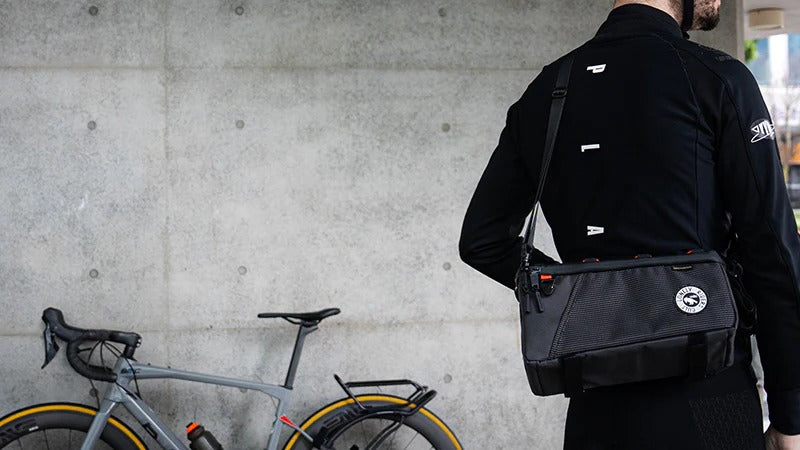 Person wearing a black bicycle pannier bag with shoulder strap with a bicycle in the foreground against a concrete wall.