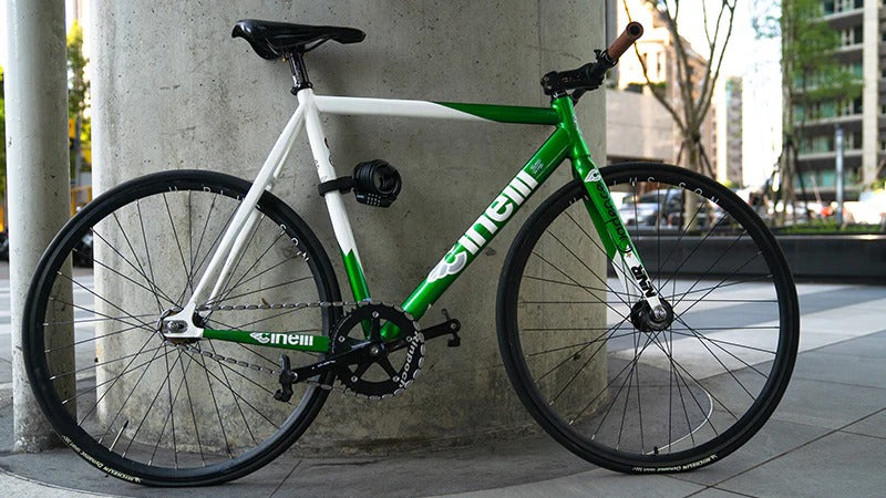 Green and white bicycle leaning against a concrete pillar in an urban setting with a bicycle cable lock attached to the downtube