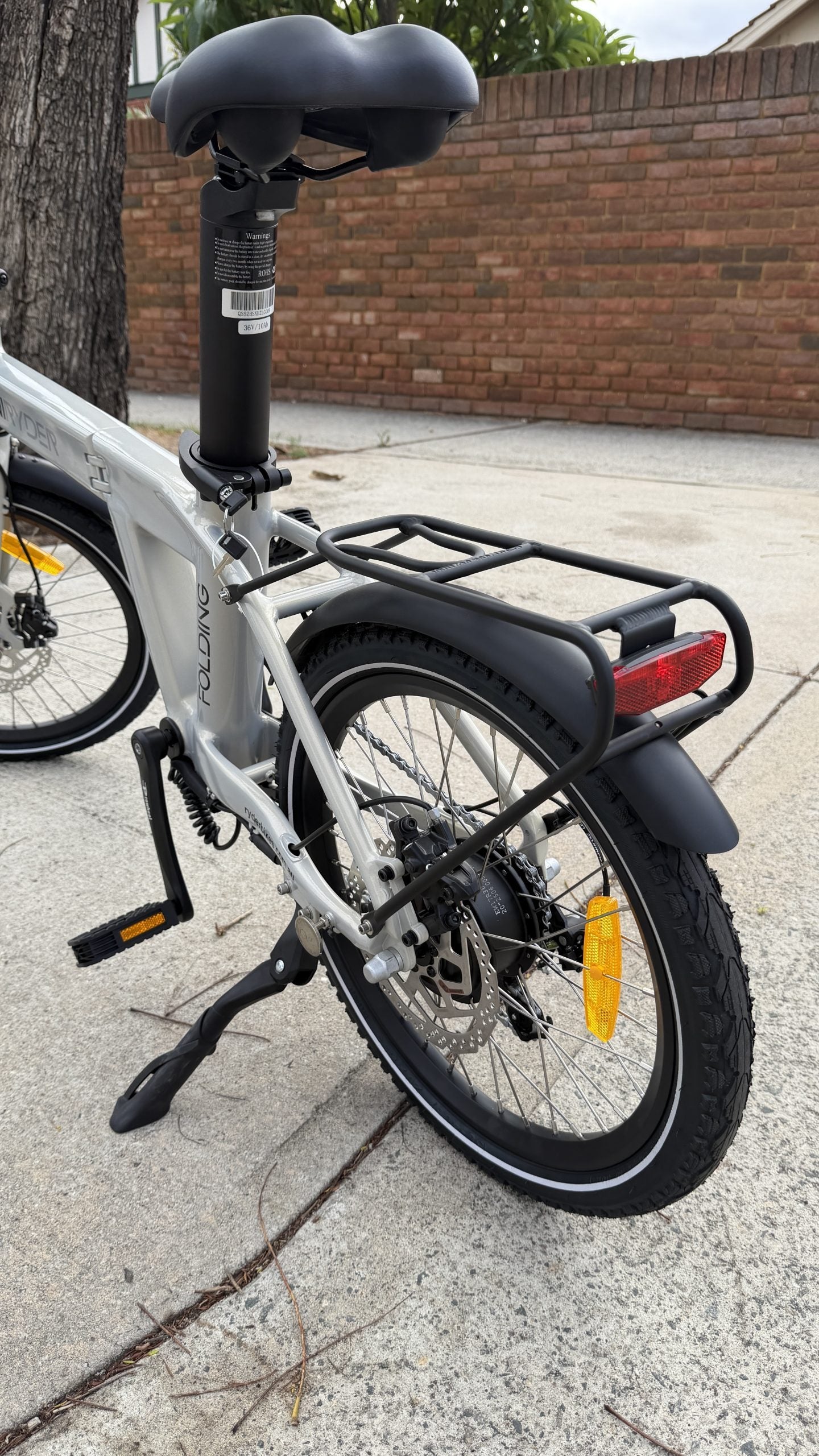 Gray electric bicycle with black seat and rear rack parked on a sidewalk next to a brick wall.