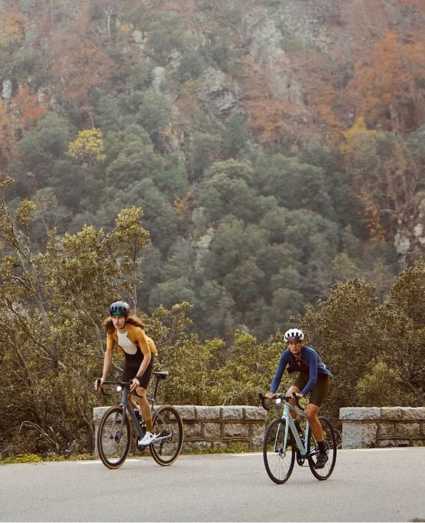 Two cyclists riding on a road with a scenic background of trees and mountains.