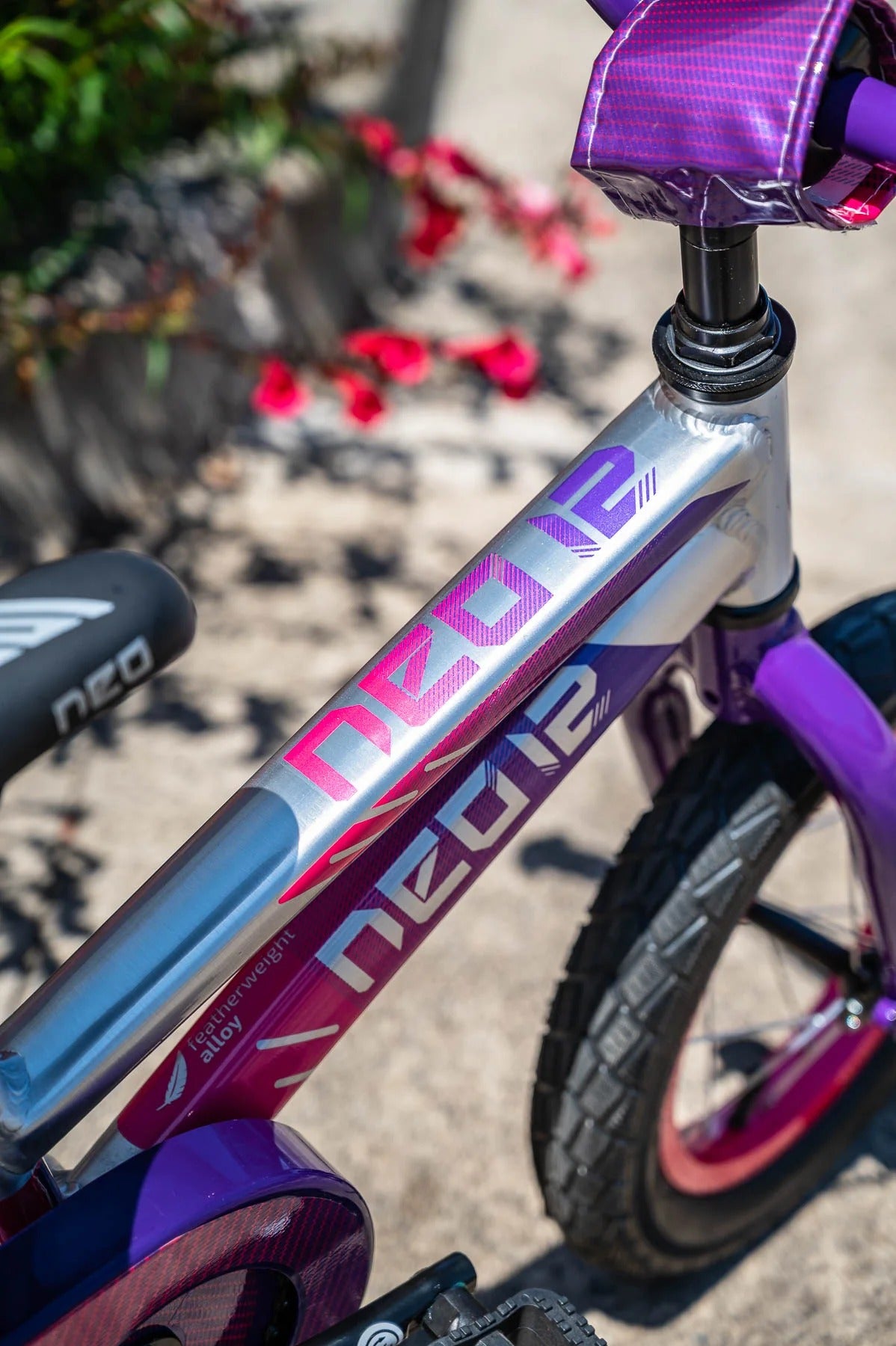 Close-up of a children's bicycle frame with a metallic silver and purple Neo brand frame, purple seat and handlebar, and a blurred outdoor background with red flowers.