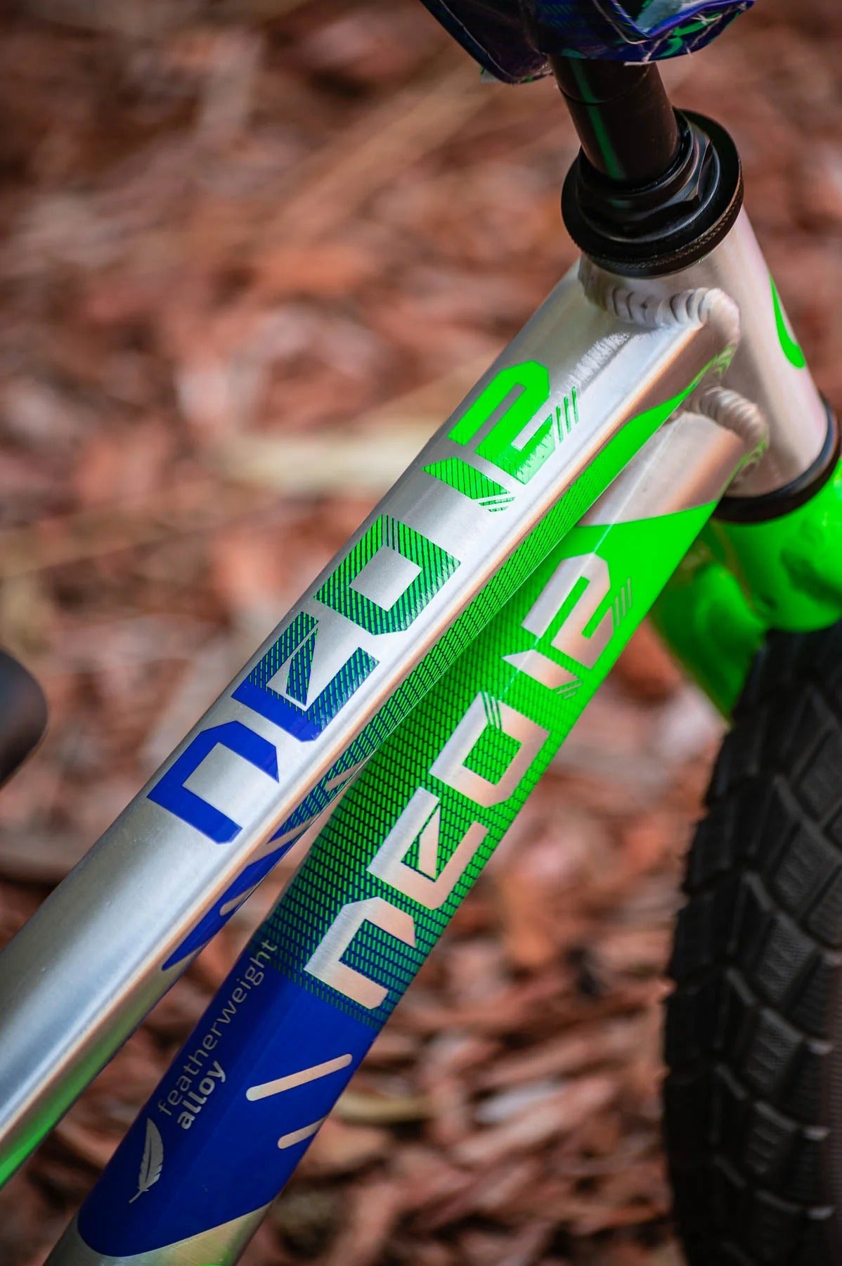 Close-up of a silver, blue and green bike frame with visible 'NEO' branding on a blurred forest floor background.