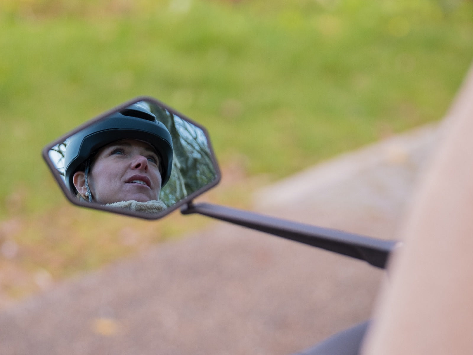 Person wearing a helmet reflected in a bicycle mirror with a blurred natural background