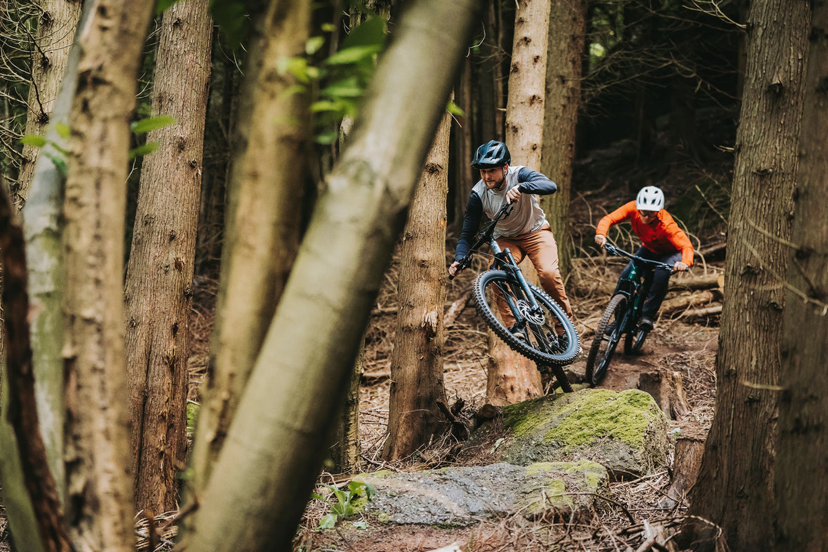 Two guys riding on mountain bikes through a forest over large boulders