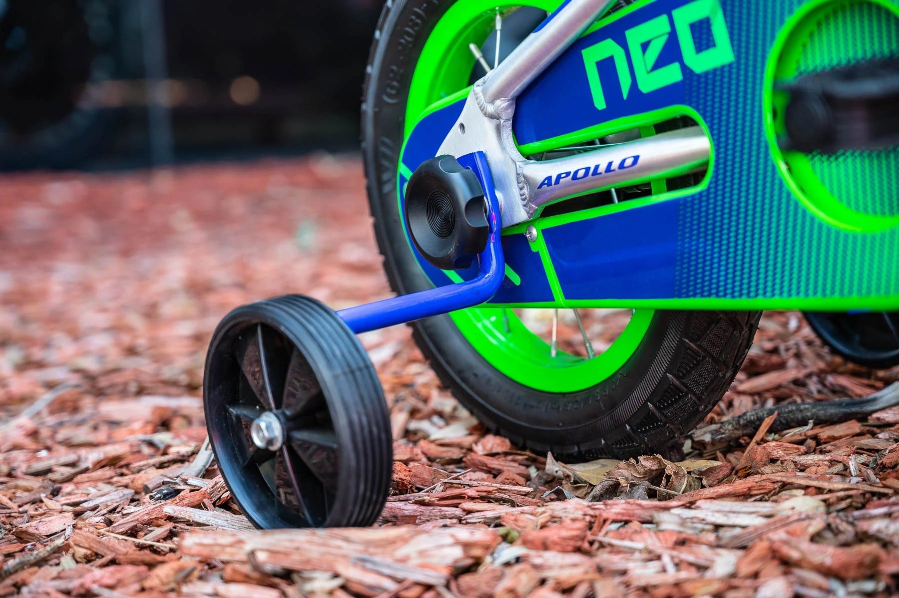 Close-up of a children's Neo bicycle wheel and training wheel on a mulch ground, showing a blue frame, bright green wheel rim, and black tyres.