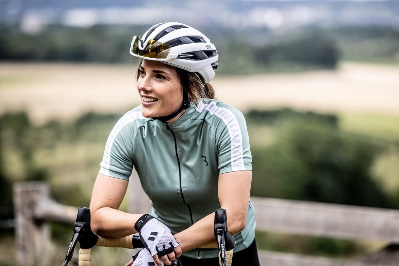 Woman in cycling gear with helmet and gloves, sitting on a bike outdoors.