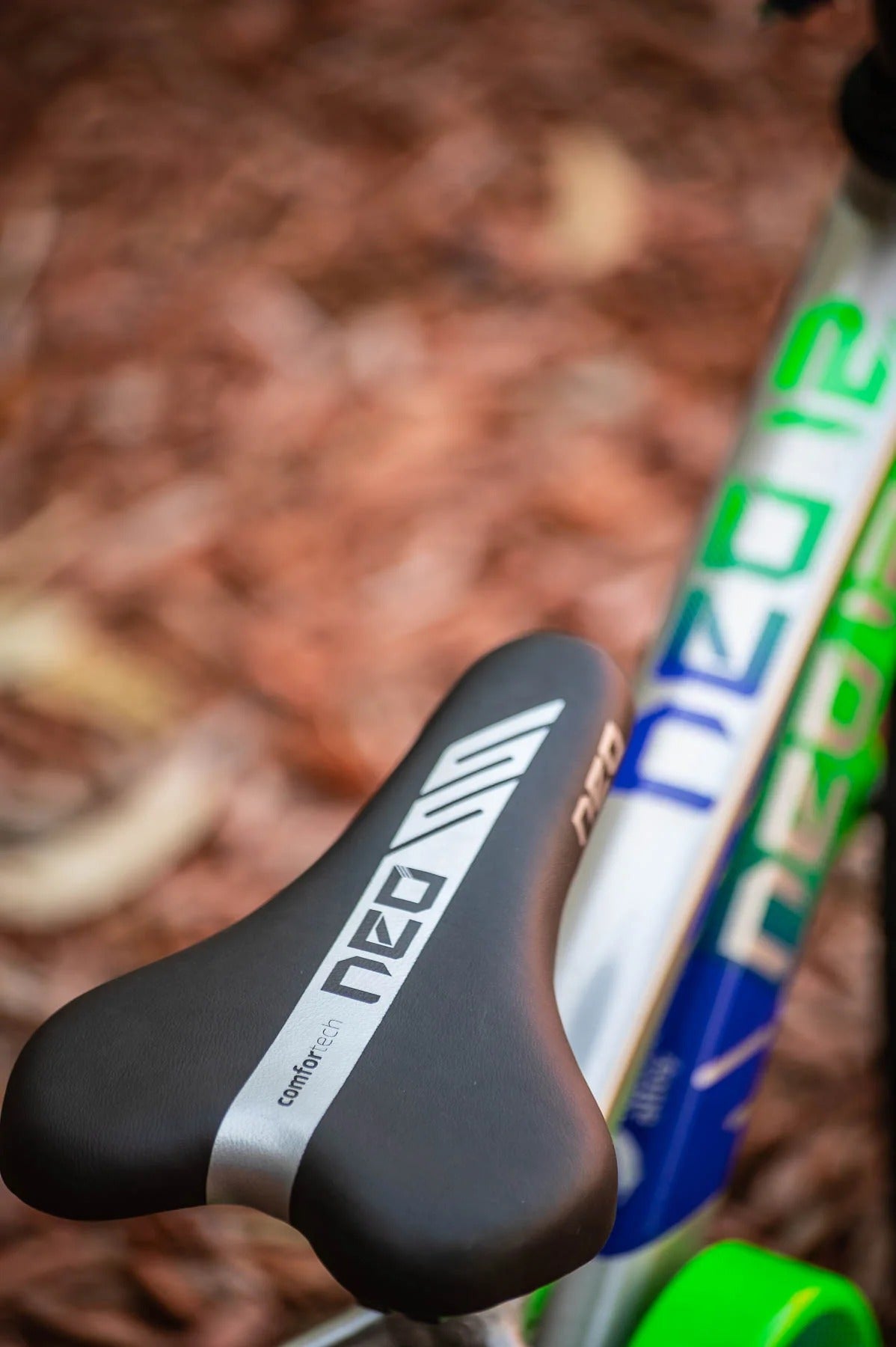 Close-up of a black Neo Pro bicycle saddle mounted on a silver blue and green frame, photographed outdoors on a blurred woodchip ground.
