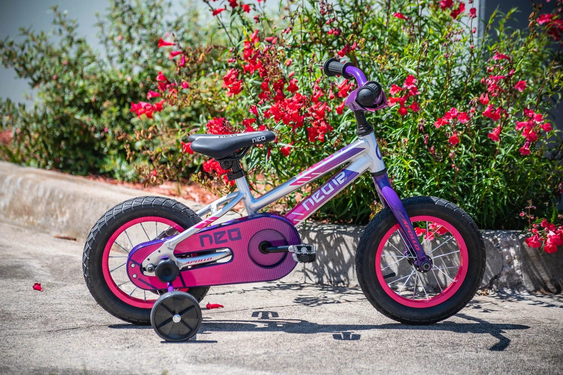 A children's Neo bicycle with training wheels, featuring a purple and silver frame with pink accents, parked on a paved path with red flowers in the background.