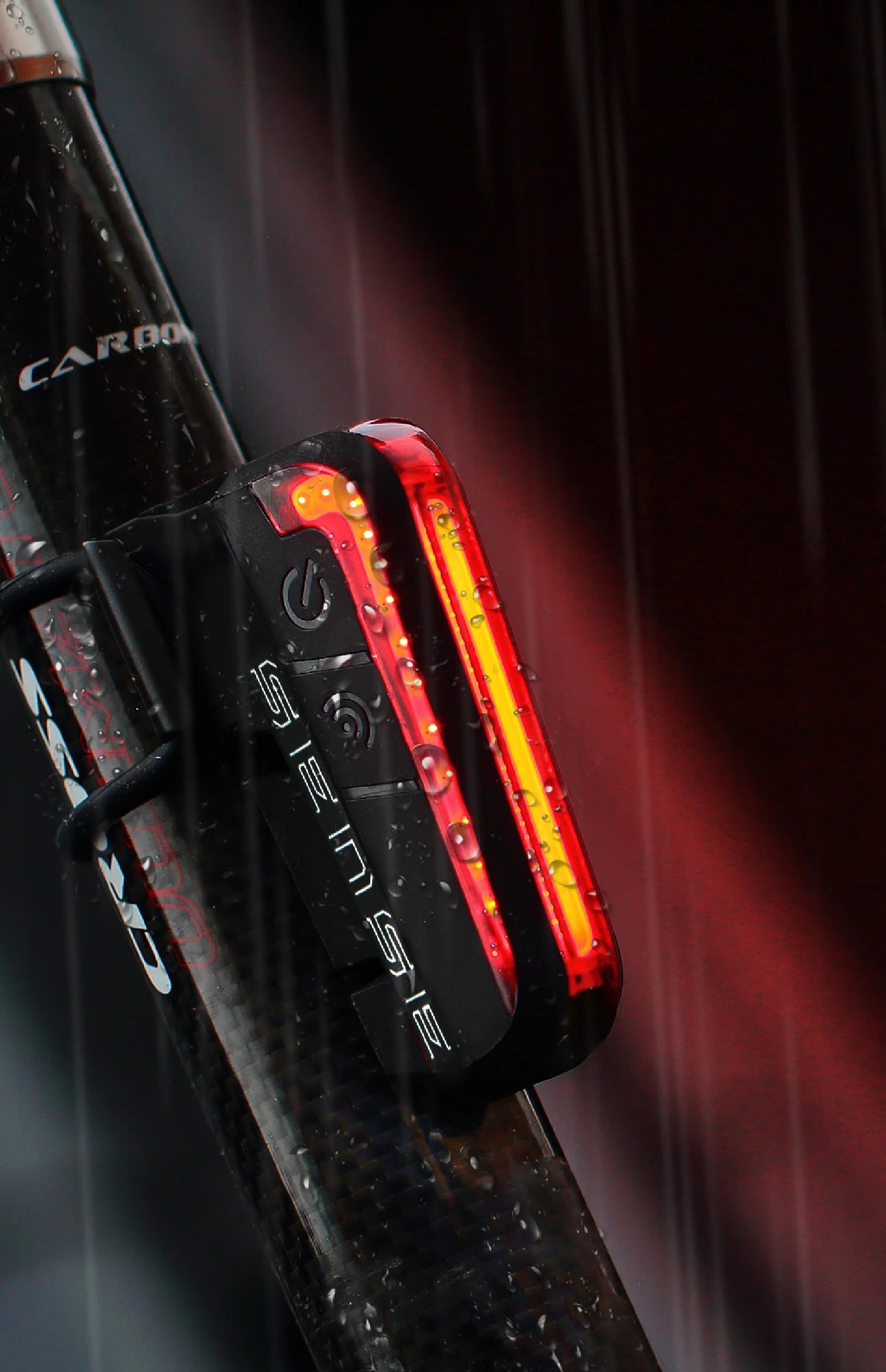 A close-up of a black bicycle rear light with a glowing red LED strip, mounted on a carbon fiber frame in the rain.