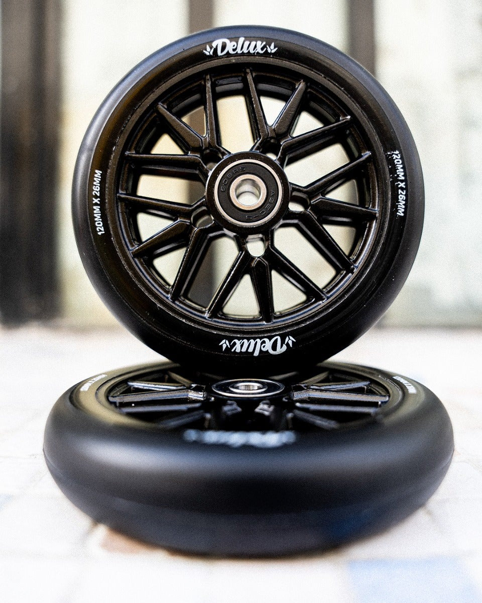Two black Delux scooter wheels stacked vertically, with the top wheel standing upright to show the multi-spoke hub and sidewall branding.