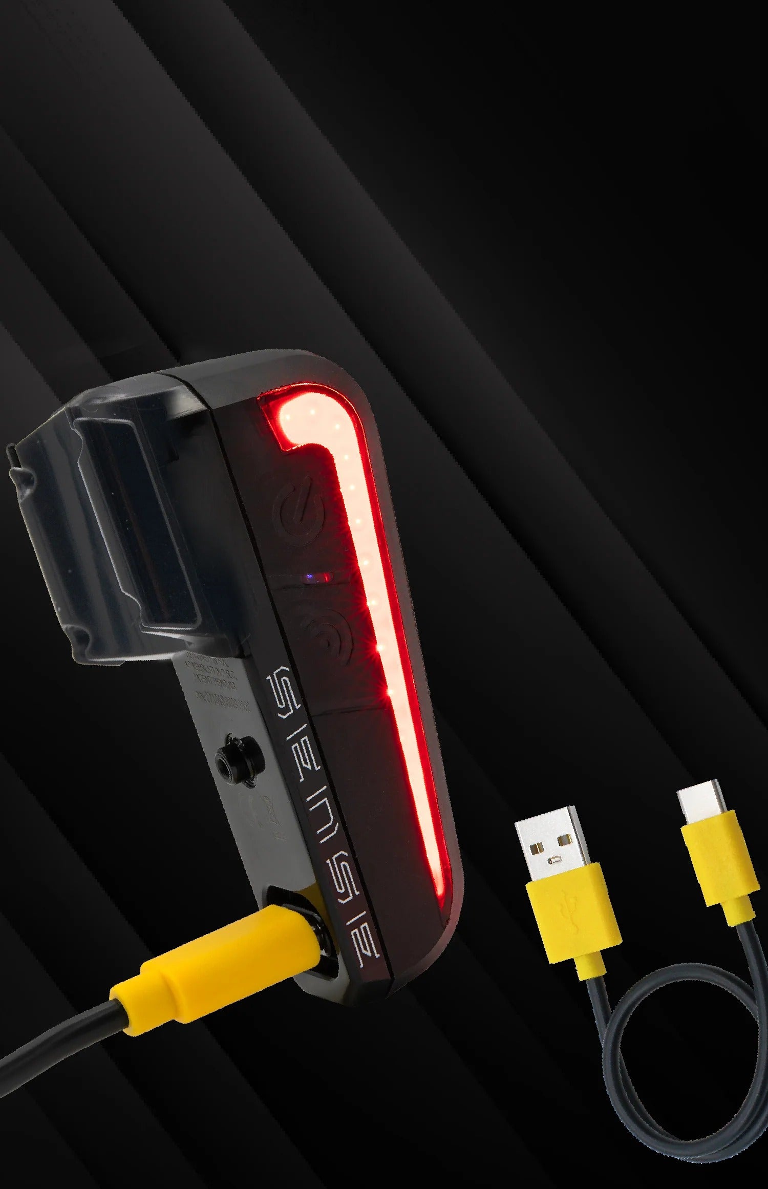 A black bicycle tail light with a glowing red LED light bar, shown with a yellow USB charging cable on a dark background. The model name 'SENSE' is visible on the side of the light.