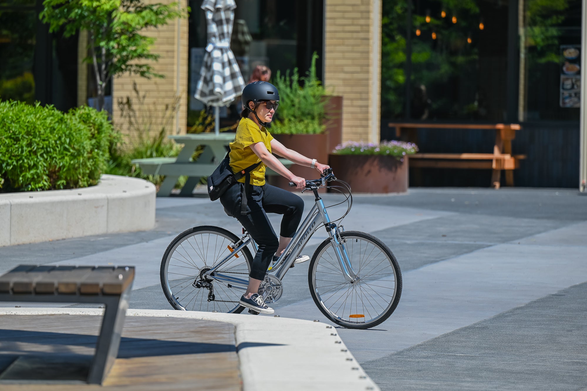 A women riding an Apollo step through hybrid bicycle on a sidewalk with a building and greenery in the background