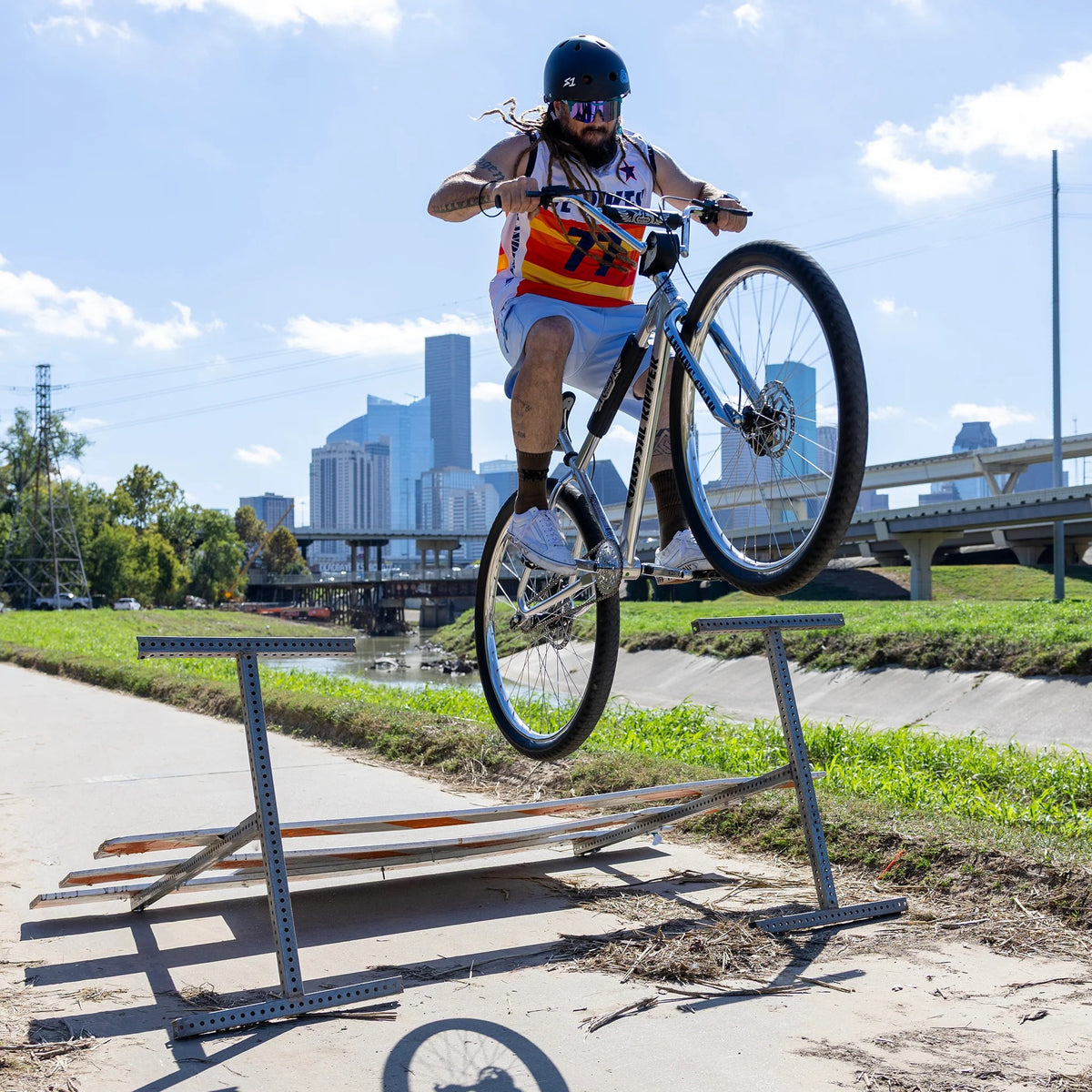 A guy jumping a silver street bicycle over a fallen street sign with a city in the background