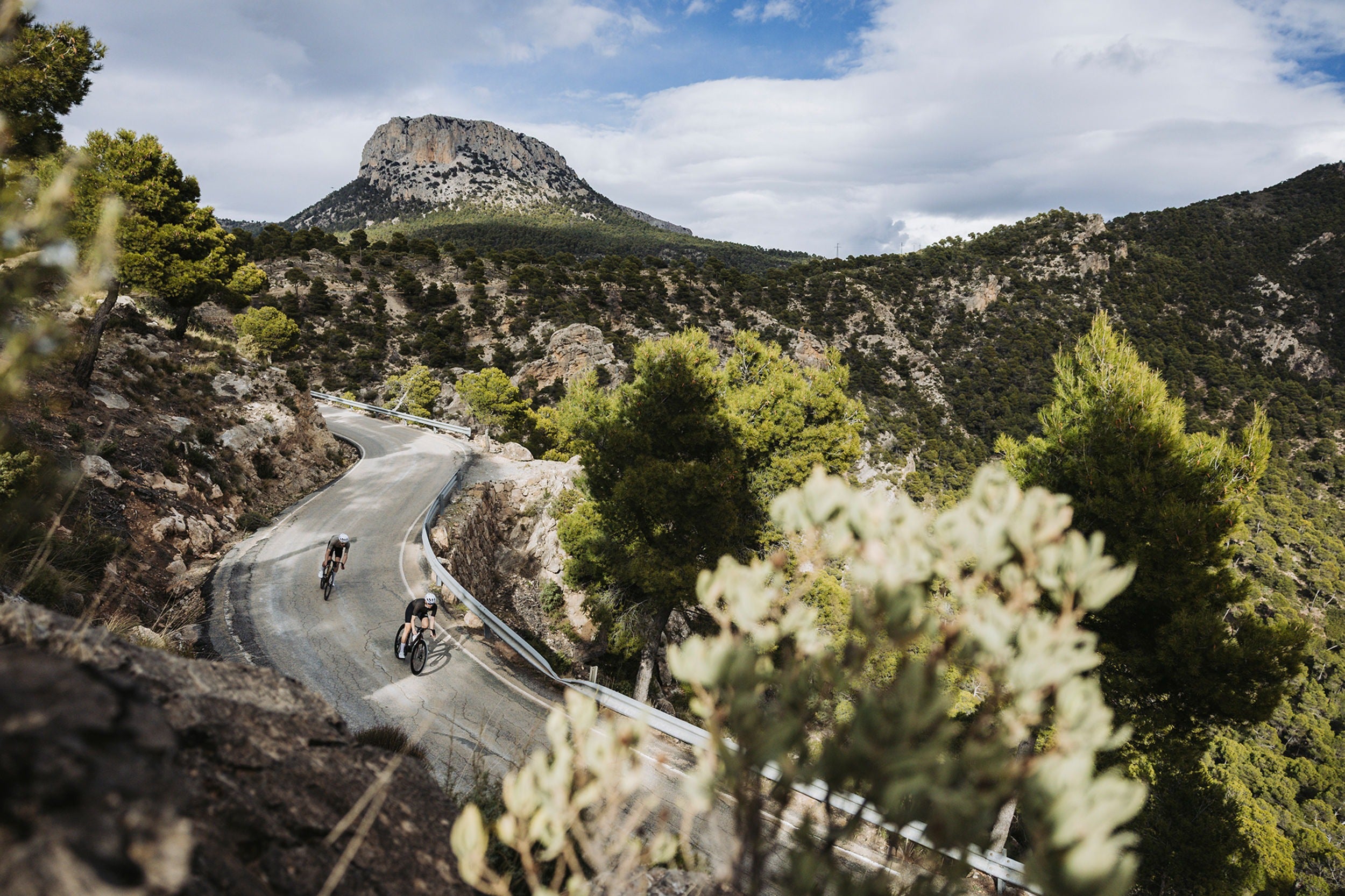 Two cyclists on a winding mountain road with a large rock formation in the background.