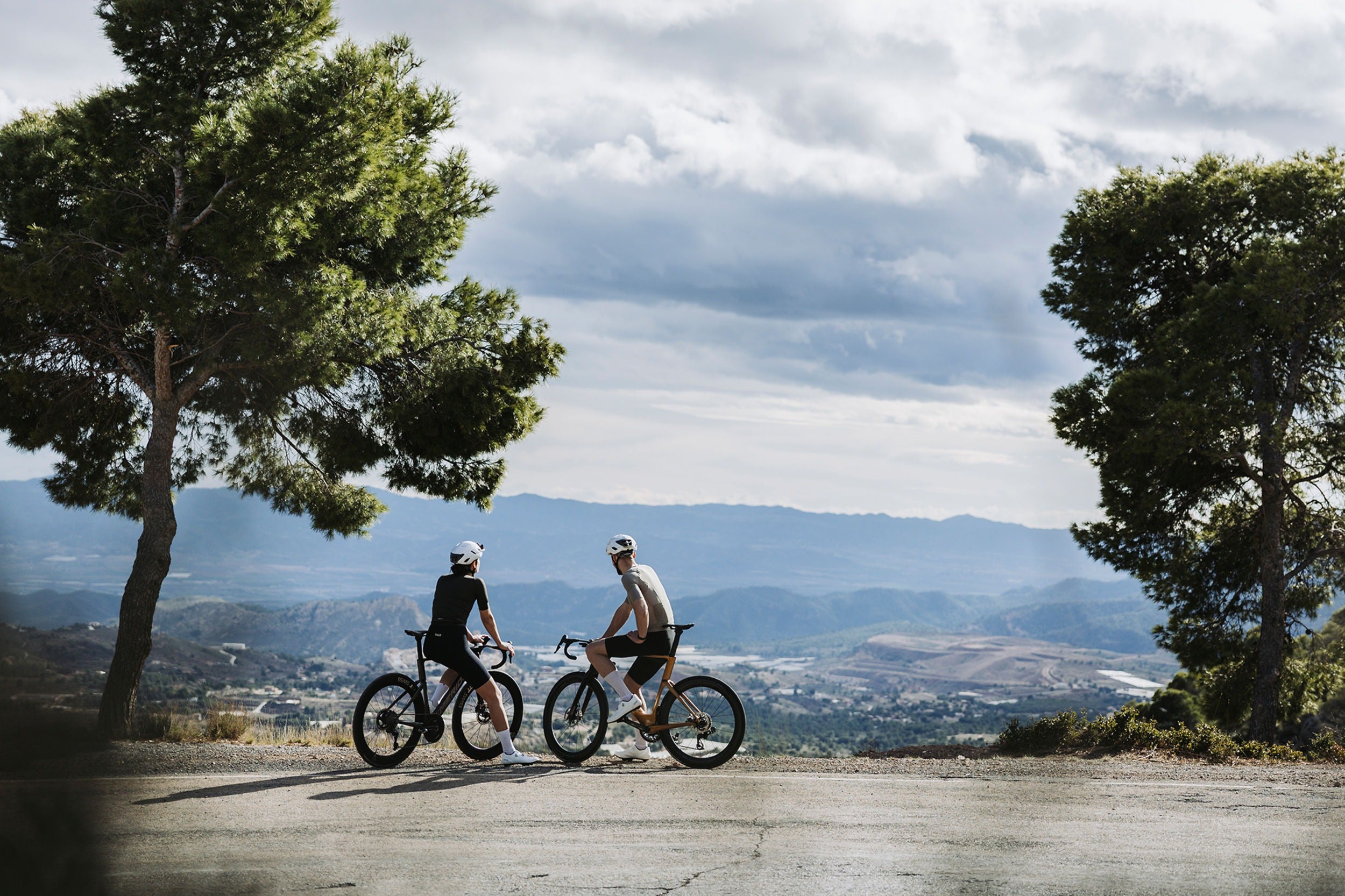 Two people riding road bicycles on a scenic road with trees and mountains in the background.
