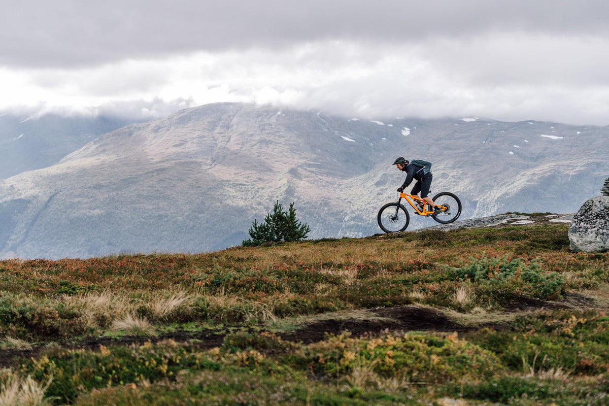 A mountain biker riding an orange dual suspension mountain bike on a trail with mountains in the background