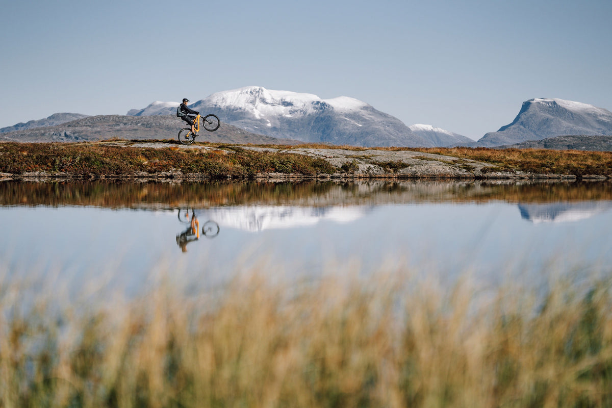 A mountain biker performing a wheelie on an orange mountain bike next to a lake with mountains in the background