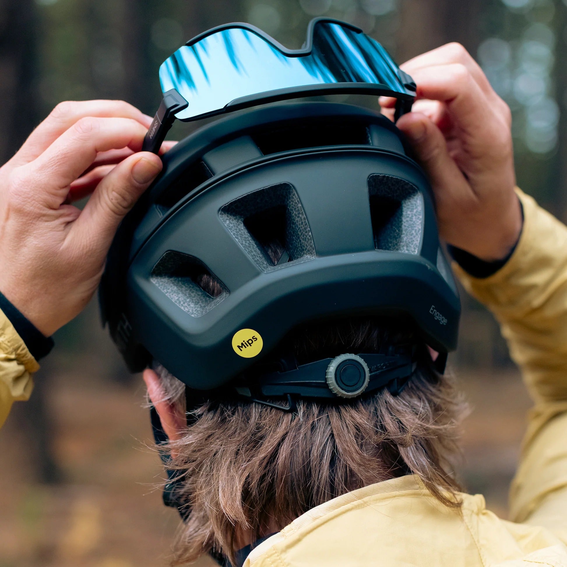The back of a person wearing a black mountain bike helmet fitting sunglasses into the back of the helmet with a forest background