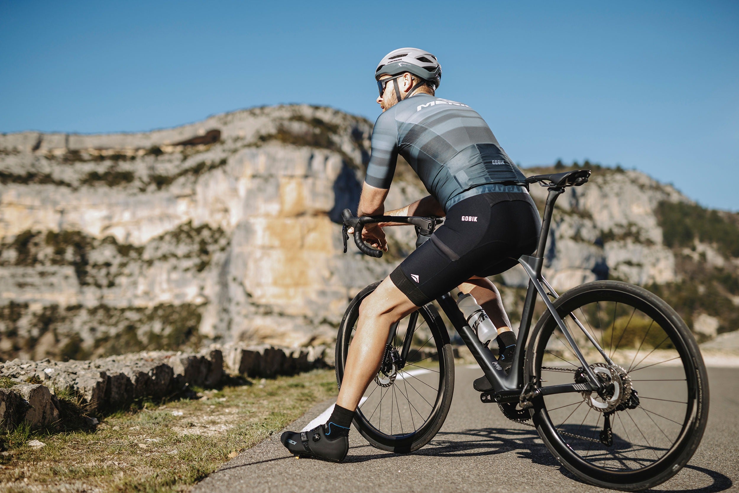 Cyclist sitting on the top tube of a road bike on a mountain road wearing a helmet and cycling outfit.