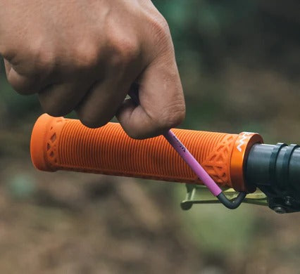 A close-up of a hand using a pin hex key to loosen a hex nut on an orange textured bicycle handlebar grip, with a blurred outdoor background.