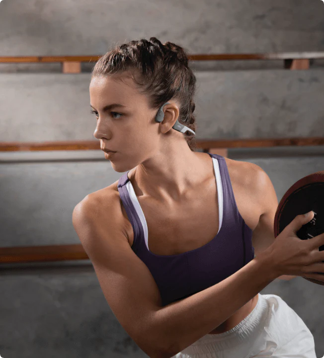Woman exercising with a weight plate, wearing wireless earbuds in a gym setting.