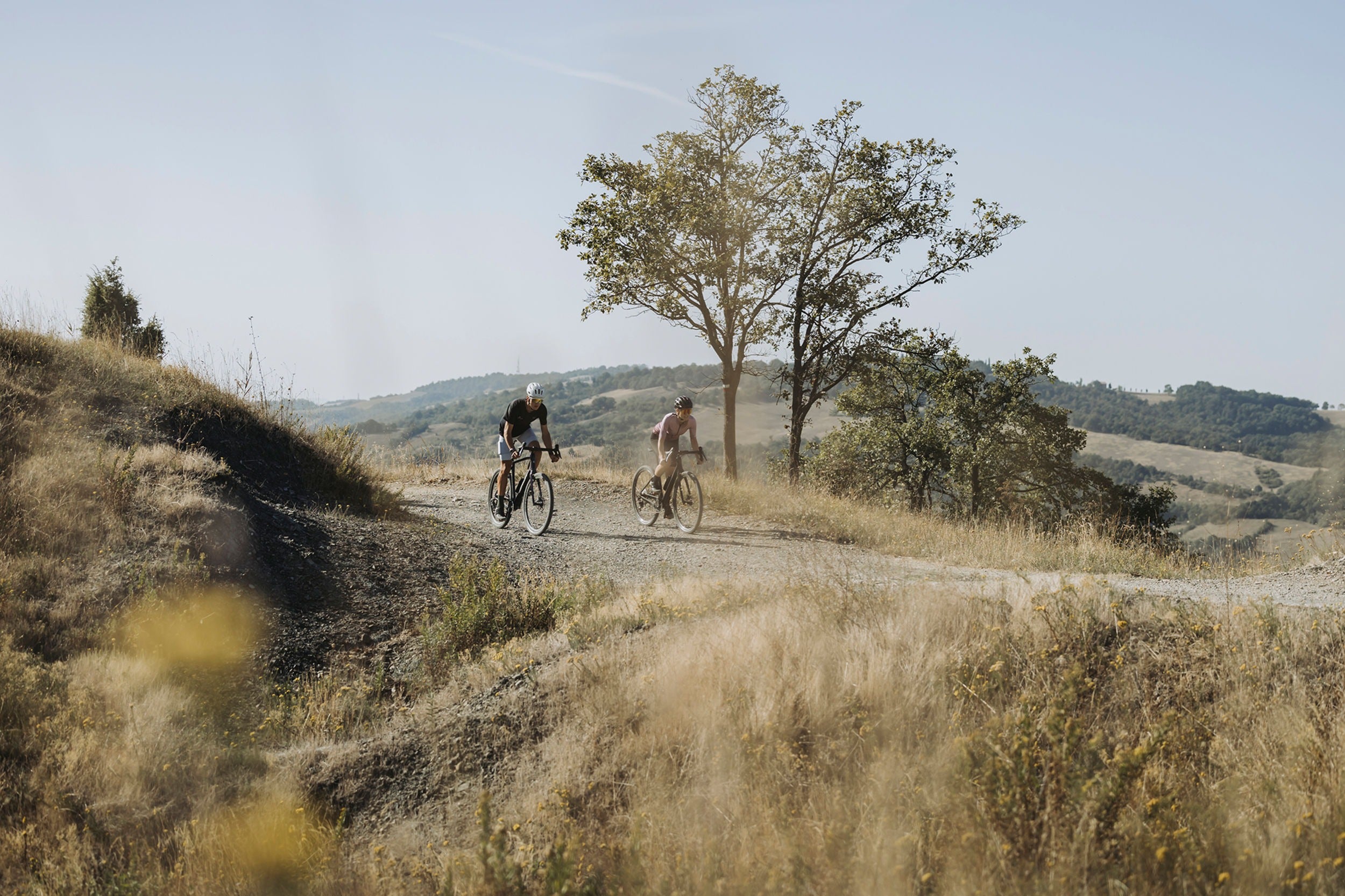 Two gravel cyclists on a dirt path with trees and open sky in the background