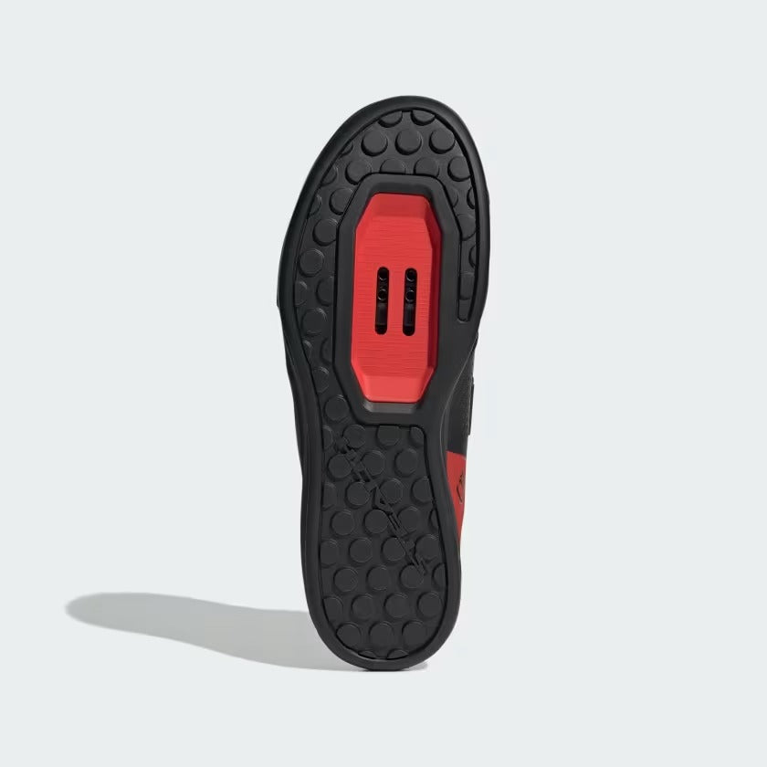The bottom sole of a cycling shoe shown upright against a light gray background, featuring a black textured tread with a prominent red central platform.