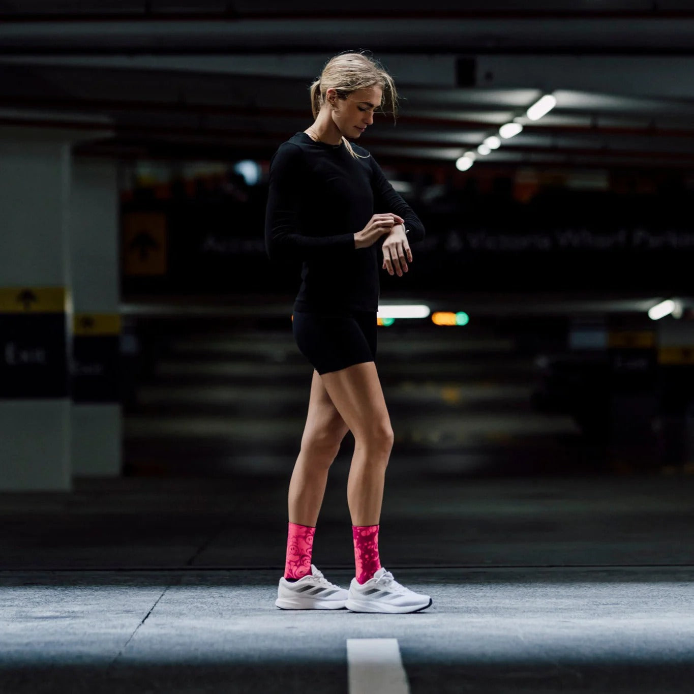 A women wearing a black gym outfitt with bright pink socks and white trainers with a blurred underground parking in the background
