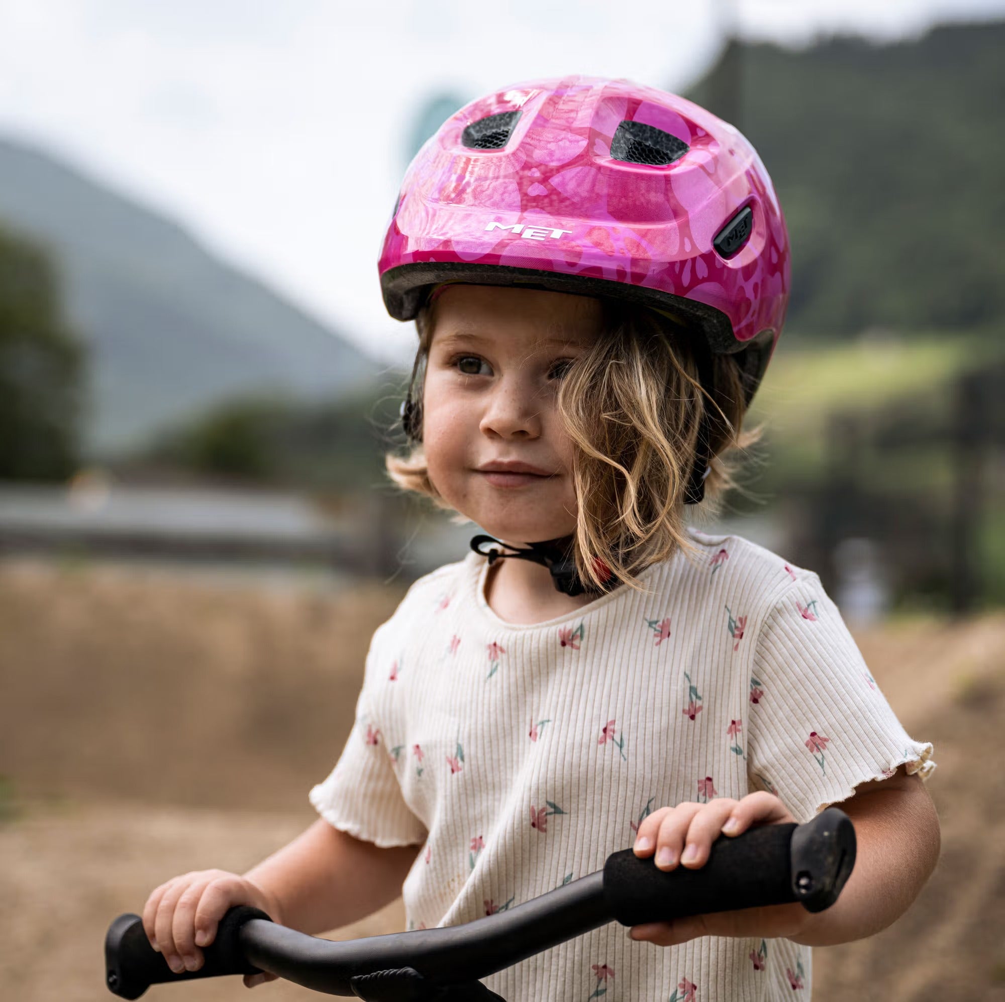 Child wearing a pink helmet and holding a scooter outdoors with mountains in the background