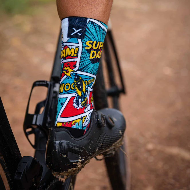 A close-up of a cyclist's lower leg wearing a black cycling shoe and a crew sock with a colorful comic book style print featuring blue, red, yellow, and white panels with text like 'SUPER DAD' and 'WOOSH!'. The background shows a blurred dirt path and bicycle frame.