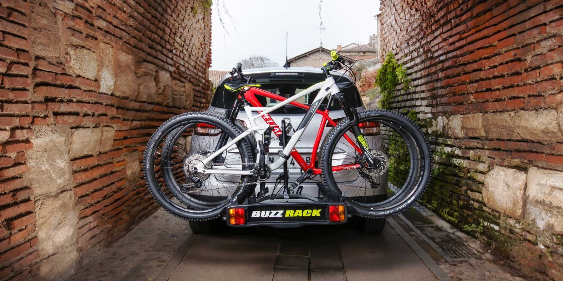 Two bicycles on a Buzz Rack attached to a car in a narrow alleyway.