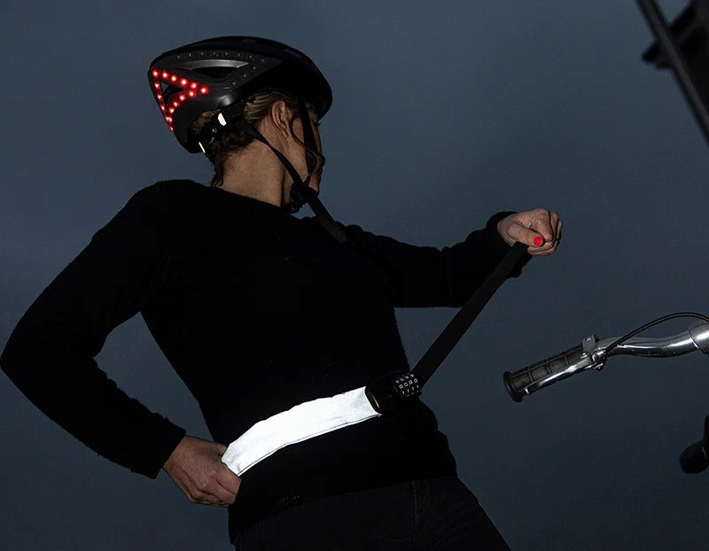 Someone wearing a bicycle helmet and securing a bicycle combo chain lock around the waist standing next to a bicycle with a dark grey background