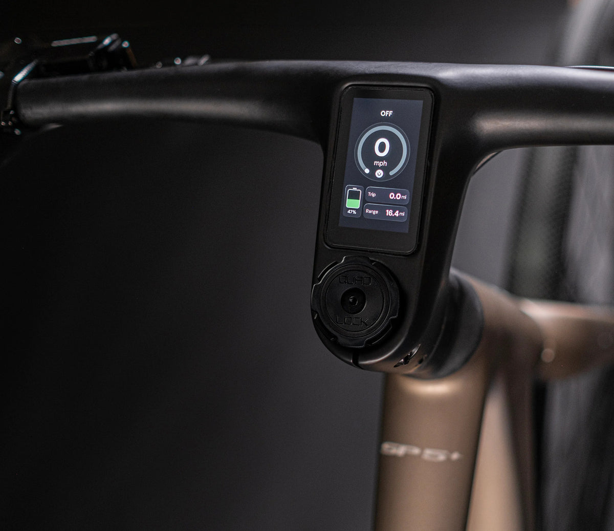 Close-up of a bicycle handlebar with an integrated black digital display screen showing cycling metrics, mounted on a metallic bronze frame.