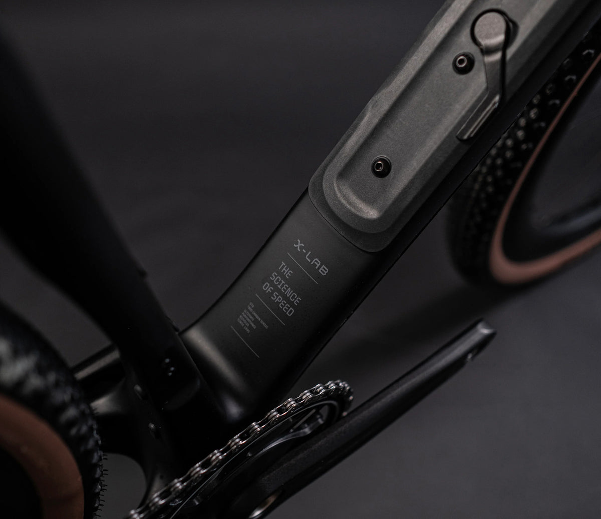 Close-up of a matte black bicycle frame section with visible text reading 'X-LAB', alongside part of the wheel and chainstay on a dark studio background.