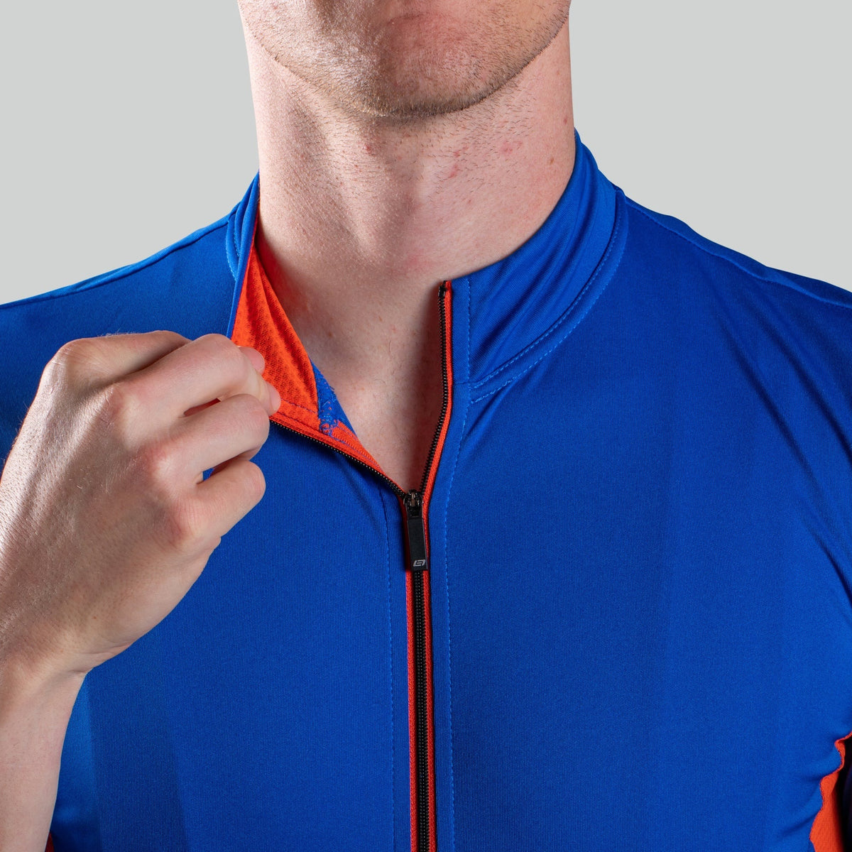 A close up of a man wearing a blue cycling jersey and peeling back the collar showing the orange on the inside of the collar on a light grey background
